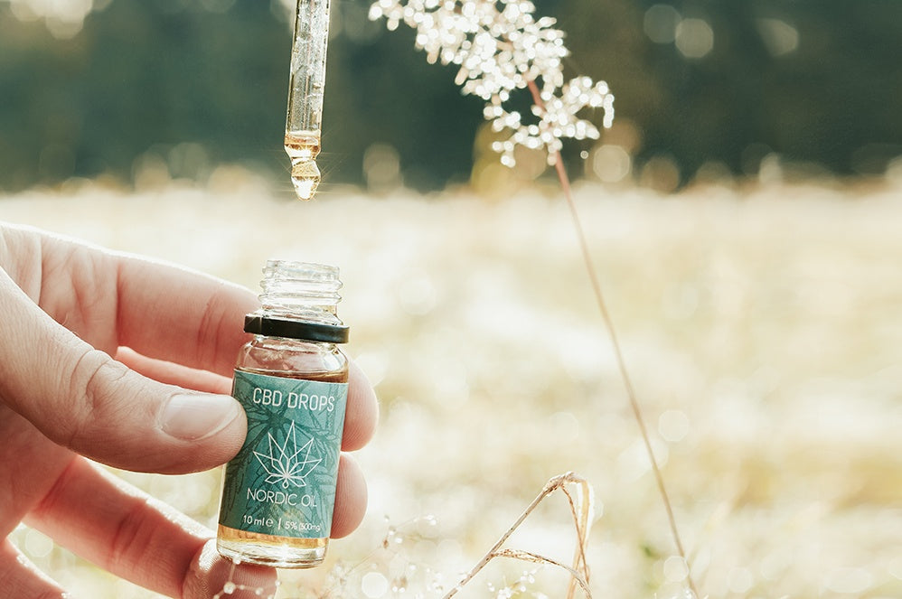 Pipette laissant tomber une goutte d'huile de CBD dans un flacon ouvert, avec un champ de blé en fond.