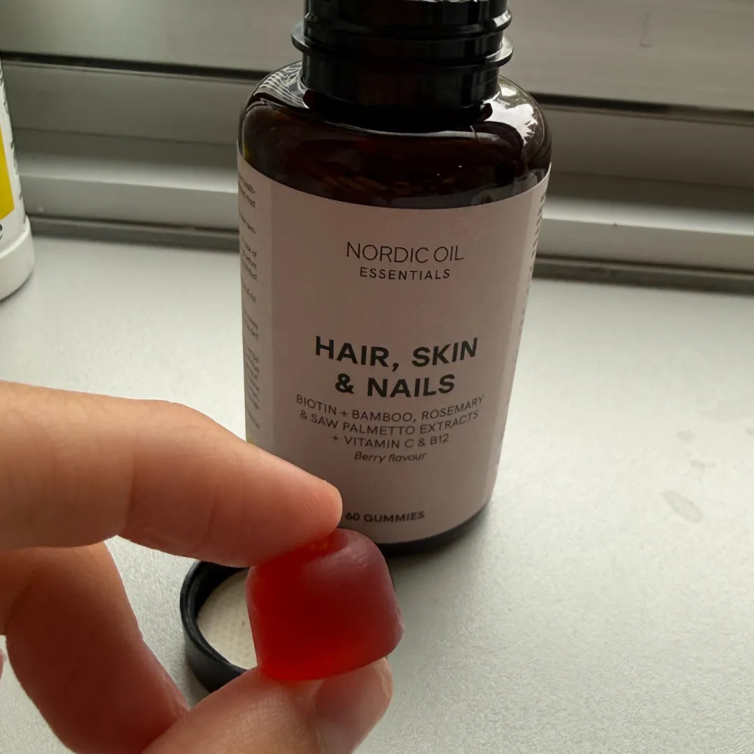 Gomme rouge tenue entre les doigts devant un flacon Nordic Oil Hair, Skin & Nails Gummies saveur fruits rouges.